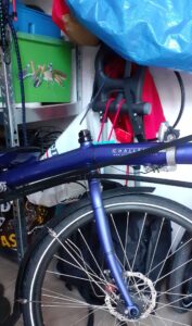 Voorwiel en een deel van het blauwe frame van een ligfiets, het merk 'Challenge' is deels te lezen. De fiets staat in een rommelige berging met kleurige spullen. 