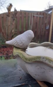 Een wit vogeltje van keramiek zit op de rand van een witte vogeldrinkschaal, tot de rand gevuld met water. Op de achtergrond een oude bruine schutting. 