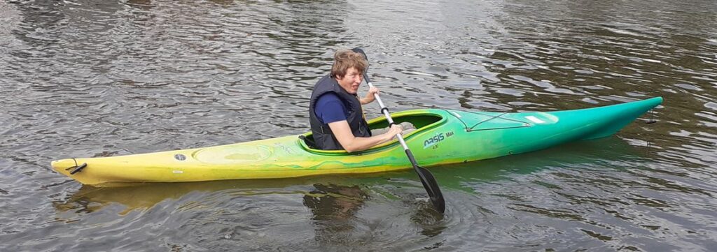 Jessica in een geel-groene kano op rustig water. Ze zit wat ongemakkelijk. 
