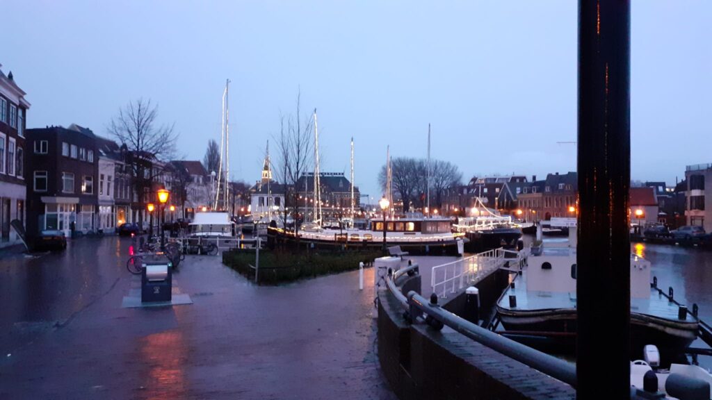 De haven in de binnenstad van Leiden met boten, versierd met lichtjes. Op de achtergrond het oude havengebouw. Het is schemerig en de straat glanst omdat de stenen nat zijn. 