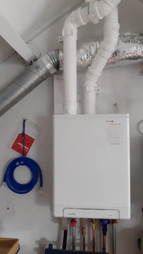 CV ketel, het is een witte doos van ca 40 cm breed en 70 cm hoog. Aan de onderkant komen er zes dunne buizen uit en aan de boven kant twee dikke witte buizen die naar het dak gaan. Links ervan hangt een opgerolde vulslang, zo dik als een tuinslang met koppelingen eraan.
