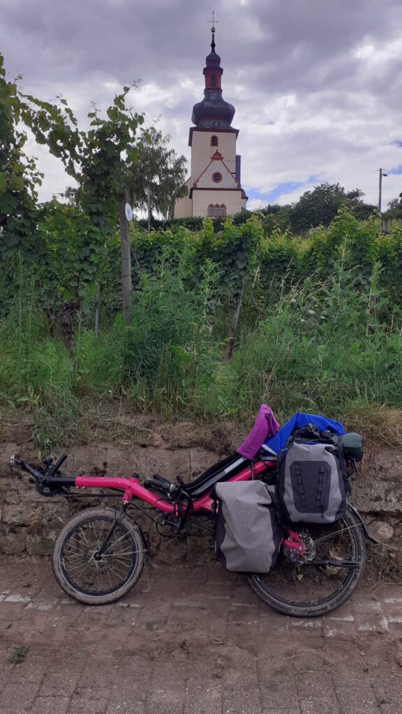 roze ligfiets met dubbele set tassen, modder aan de wielen. daarachter allerlei groen en een kerkje met een toren in de vorm van een ui